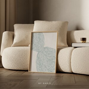 May include: A framed abstract art print with a light blue and white design. The print is in a wooden frame and is sitting on a white couch.
