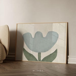 May include: Abstract art print in a wooden frame, depicting a large blue flower with green leaves on a neutral backdrop. The artwork is leaning against a wall. The text "BY GABIS" is visible at the bottom of the print.