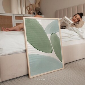 May include: Abstract art print featuring green and blue shapes on a white background. The print is framed in a light wood frame and is leaning against a white bed.