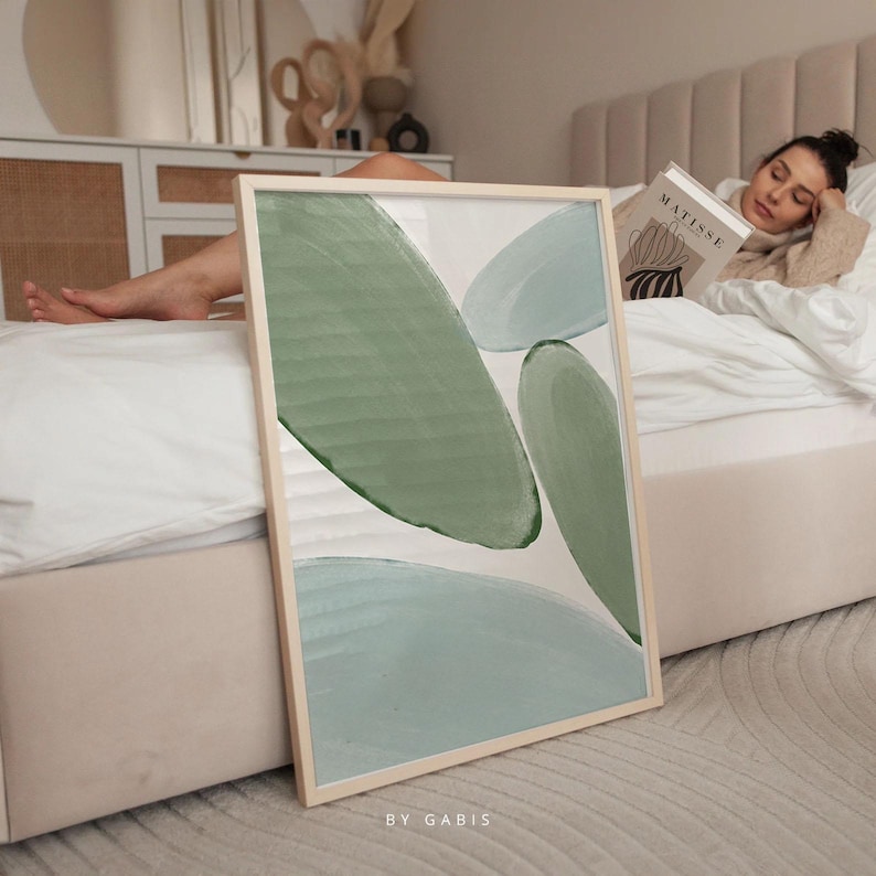 May include: Abstract art print featuring green and blue shapes on a white background. The print is framed in a light wood frame and is leaning against a white bed.