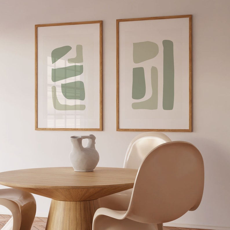 May include: Two framed abstract art prints with a green and white color scheme. The prints are hanging on a white wall above a wooden table and two chairs.