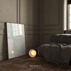 May include: A gray couch with a throw blanket in a room with a framed abstract art print and a round, gold table lamp with a white shade. The room has dark wood floors and white walls with decorative molding.