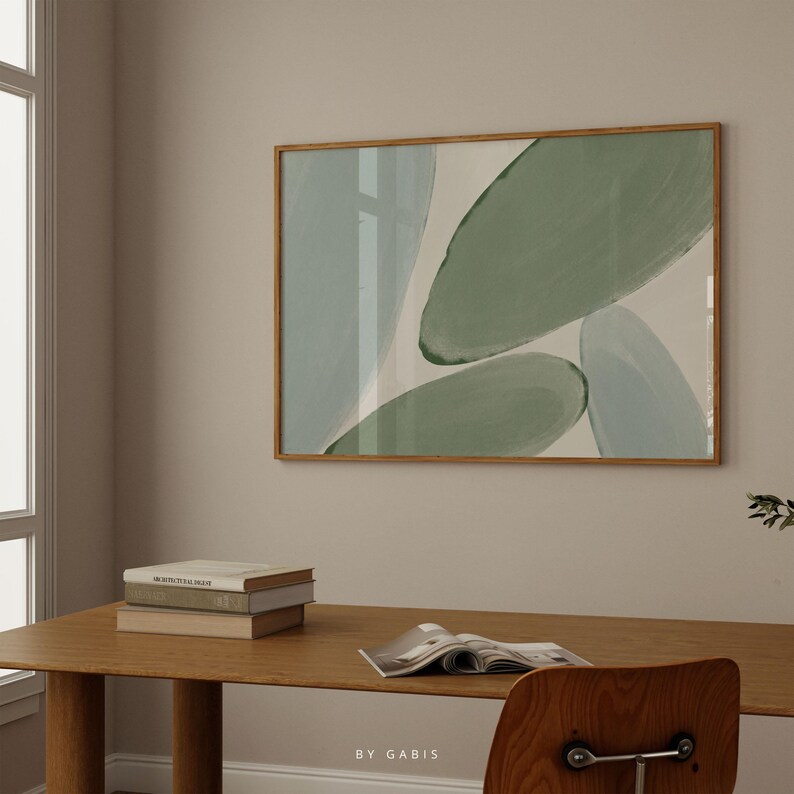 May include: A framed abstract painting with green and blue shapes on a white background. The painting is hanging on a beige wall above a wooden desk with books and a magazine.