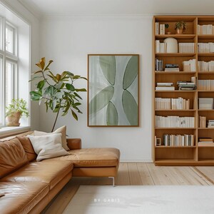 May include: A light brown leather sectional sofa with a white throw pillow in a living room with a large abstract painting of green leaves and a wooden bookshelf filled with books.