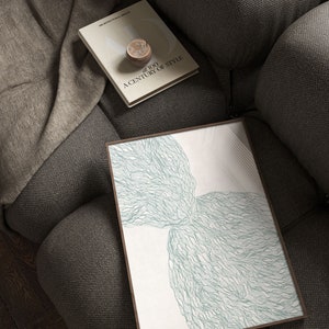 May include: A framed abstract art print with a white background and a green, organic, flowing design. The print is on a gray couch with a book titled "A Century of Style" on top of it.