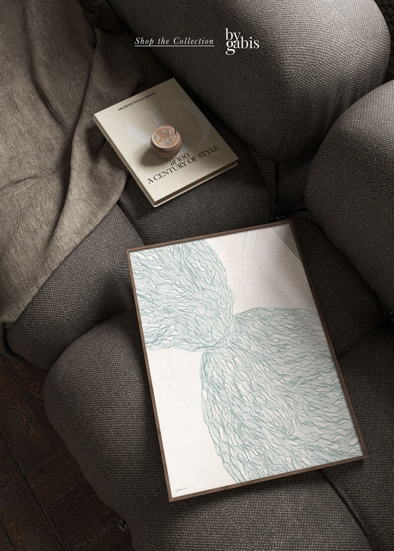 May include: A framed abstract art print with a white background and a green, organic, flowing design. The print is on a gray couch with a book titled "A Century of Style" on top of it.