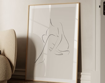 May include: A framed art print featuring a minimalist line drawing of a seated figure. The artwork is rendered in black on a neutral beige background, encased in a light wood frame. The print is displayed against a light-colored wall, with a glimpse of a chair and wooden floor.