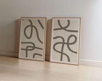 Puede incluir: Dos impresiones artísticas abstractas enmarcadas. Cada impresión presenta un fondo beige con un diseño de línea continua gris oscuro. Los marcos son de color madera clara. Las impresiones están apoyadas contra una pared blanca sobre un suelo de madera clara. El texto "BY GABIS" está en la parte inferior.