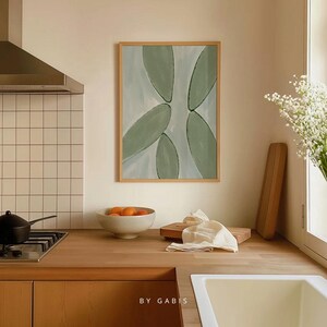 May include: A framed abstract painting with a light blue background and green shapes. The painting is hanging on a white wall above a wooden countertop.