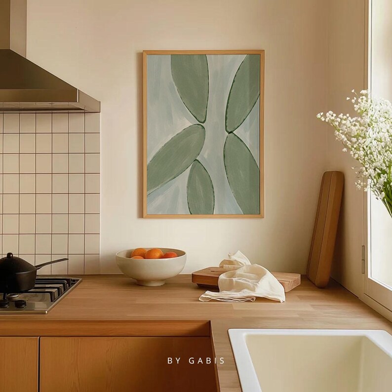 May include: A framed abstract painting with a light blue background and green shapes. The painting is hanging on a white wall above a wooden countertop.