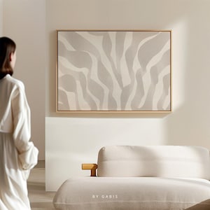 May include: A white and grey abstract painting with a zebra-like pattern. The painting is framed in a light wood frame and is hanging on a white wall. The painting is in a modern living room with a white sofa.