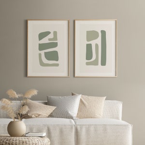 May include: Two framed abstract art prints with a light green and white color scheme. The prints are hanging on a light beige wall above a white couch with beige and white patterned pillows.