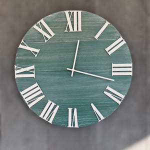 May include: A round wall clock with a teal-colored wooden face and white Roman numerals. The clock hands are white. The clock is mounted on a gray wall. The clock's diameter is approximately 12 inches.
