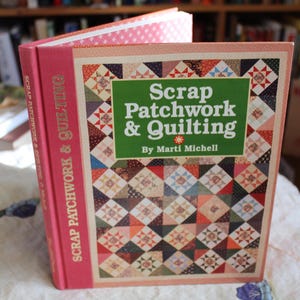 May include: A hardcover book titled "Scrap Patchwork & Quilting" by Marti Michell. The book has a pink spine with gold lettering and a cover featuring a patchwork quilt design in various colors. The book is open, revealing the patterned pages.