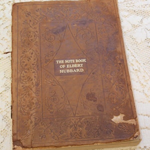 May include: A brown leather-bound book with a gold embossed title: "The Note Book of Elbert Hubbard". The cover features a decorative floral design.
