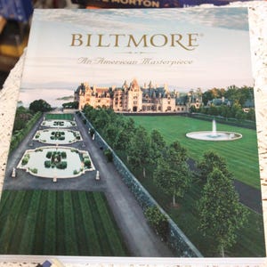 May include: A hardcover book titled "Biltmore: An American Masterpiece" featuring a scenic view of the Biltmore Estate. The cover displays the estate's architecture, gardens, and a fountain, with a backdrop of a serene sky.