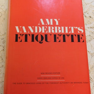 May include: A red hardcover book titled "AMY VANDERBILT'S ETIQUETTE" with the text "NEW REVISED EDITION" and "OVER 2,500,000 COPIES IN USE" on the cover. The book is subtitled "THE GUIDE TO GRACIOUS LIVING BY THE FOREMOST AUTHORITY ON MANNERS TODAY."