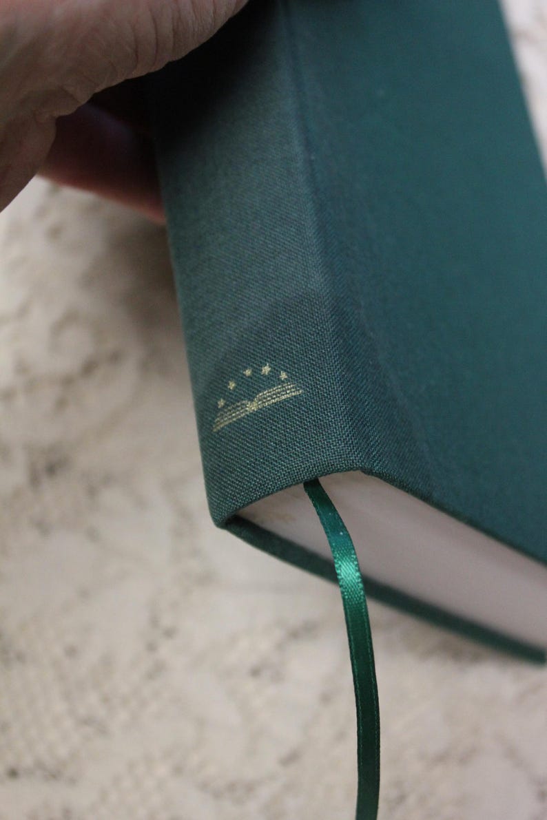 May include: A dark green hardcover book with a gold ribbon bookmark. The book has a gold embossed design on the cover that includes a book and five stars.