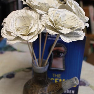 May include: A display of paper roses crafted from book pages, arranged in a small, decorative ceramic vase. The vase features a brown and grey colour scheme and a distinctive spout. A blue book is visible.