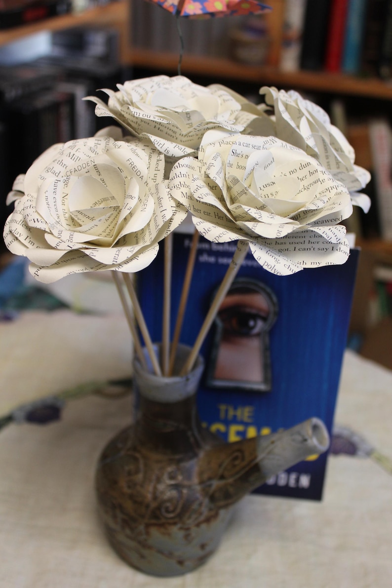 May include: A display of paper roses crafted from book pages, arranged in a small, decorative ceramic vase. The vase features a brown and grey colour scheme and a distinctive spout. A blue book is visible.