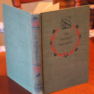 May include: A hardcover book titled The Wright Brothers with a sage green cloth cover. The front features a black illustration of an early airplane above the title, surrounded by a red and black leaf wreath. The spine includes the title and text.