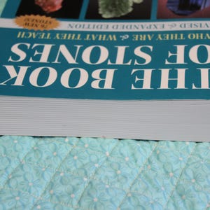 May include: A teal book titled "The Book of Stones" with white lettering. The book's cover features images of various stones and the text "Who They Are & What They Teach." The book is on a light blue quilted surface.