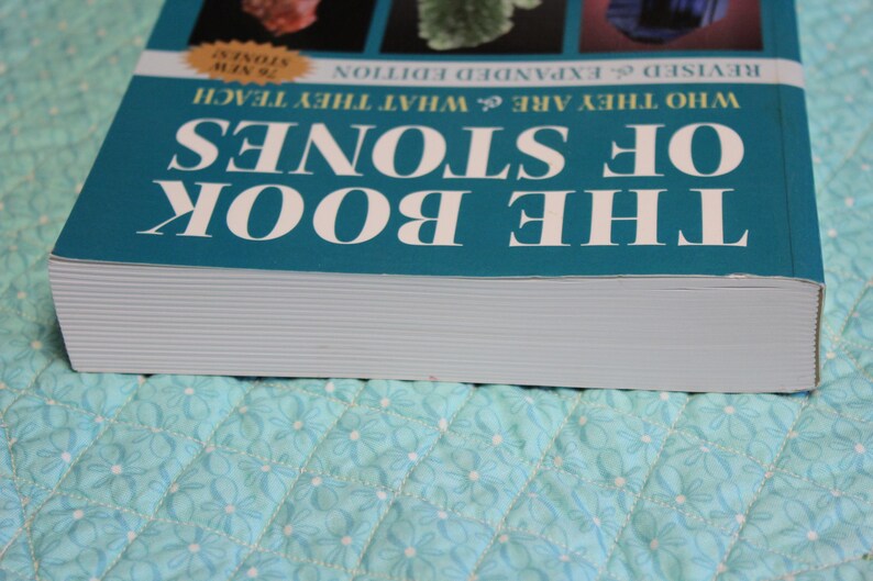 May include: A teal book titled "The Book of Stones" with white lettering. The book's cover features images of various stones and the text "Who They Are & What They Teach." The book is on a light blue quilted surface.