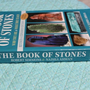 May include: A turquoise book titled "The Book of Stones" by Robert Simmons and Naisha Ahsian. The cover features images of various stones and the text "Who They Are & What They Teach". A gold sticker reads "Revised & Expanded Edition".