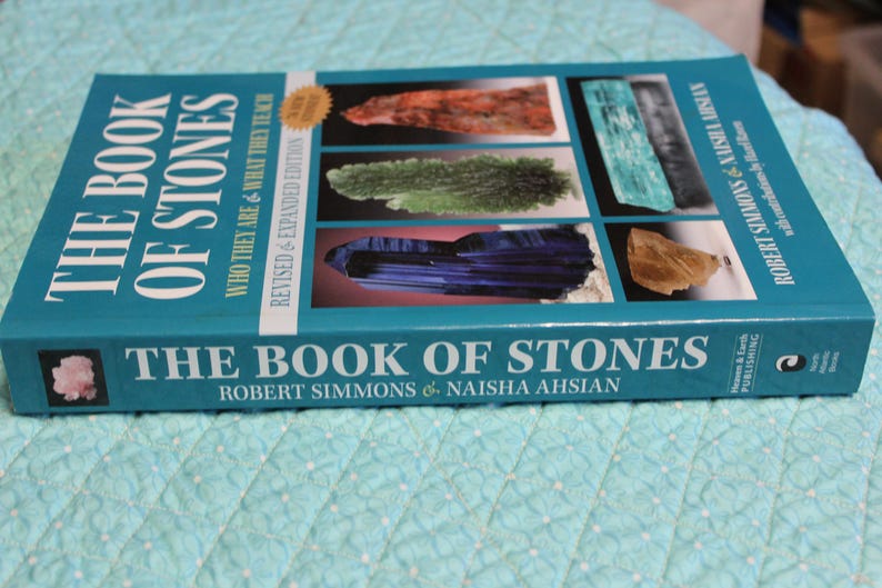 May include: A turquoise book titled "The Book of Stones" by Robert Simmons and Naisha Ahsian. The cover features images of various stones and the text "Who They Are & What They Teach". A gold sticker reads "Revised & Expanded Edition".