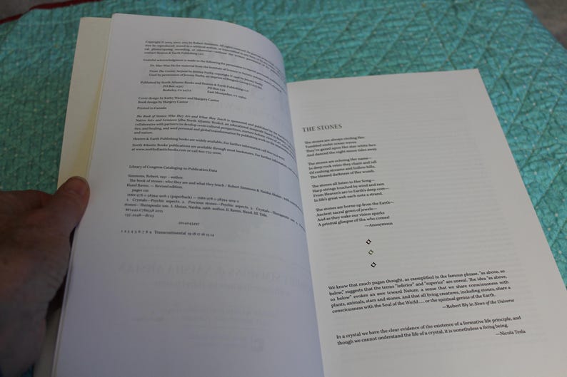 May include: An open book with white pages, featuring text on both sides. The left page displays copyright information and details, while the right page presents a poem titled "The Stones." The book is held open by a hand.