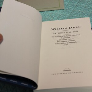 May include: A hardcover book titled William James Writings 1902-1910, featuring a white title page with black text listing works including The Varieties of Religious Experience, Pragmatism, A Pluralistic Universe, The Meaning of Truth, and Some Problems of Philosophy.
