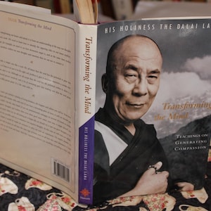 May include: An open book titled "Transforming the Mind" by His Holiness the Dalai Lama. The cover features a portrait of the Dalai Lama and the book's title. The book is open to reveal printed text on the pages.