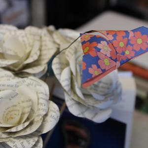 May include: A bouquet of paper roses crafted from book pages, featuring a blue and floral origami accent. The paper flowers have a vintage appearance, with visible text from the book pages. The origami is blue with pink and yellow flowers.