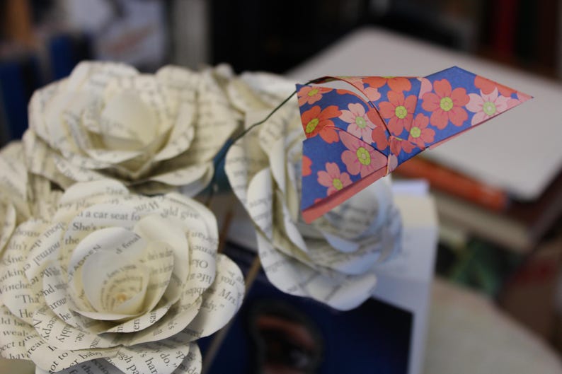 May include: A bouquet of paper roses crafted from book pages, featuring a blue and floral origami accent. The paper flowers have a vintage appearance, with visible text from the book pages. The origami is blue with pink and yellow flowers.