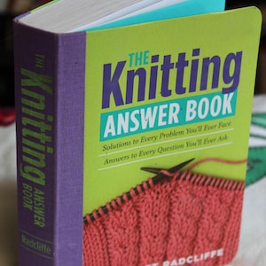 May include: A green and purple book titled "The Knitting Answer Book" by Margaret Radcliffe. The book cover features a close-up of a knitted fabric in a pink color.
