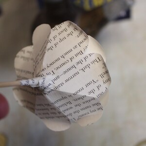 May include: A handcrafted paper flower made from book pages. The flower is white with visible text, attached to a wooden skewer. The petals are shaped to resemble a flower, a unique decorative item.