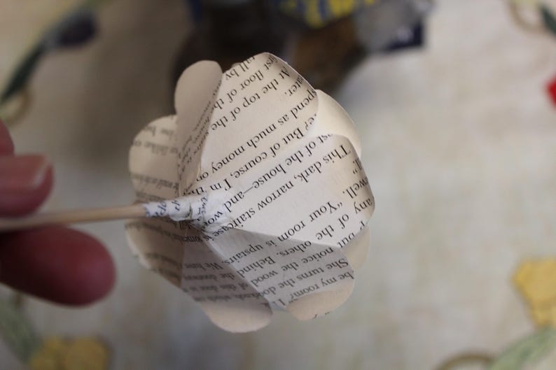 May include: A handcrafted paper flower made from book pages. The flower is white with visible text, attached to a wooden skewer. The petals are shaped to resemble a flower, a unique decorative item.