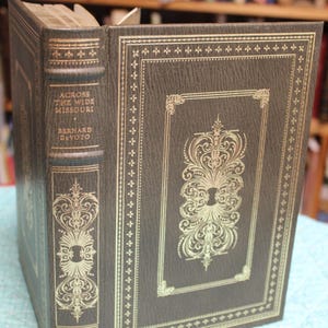 May include: A hardcover book titled "Across the Wide Missouri" by Bernard DeVoto. The book has a dark brown cover with gold decorative accents and text. The spine and cover feature ornate designs.
