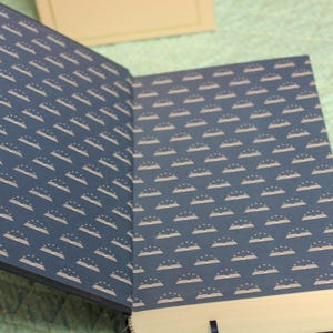 May include: An open hardcover book featuring dark blue endpapers with a repeating white pattern of open books topped with three small stars. The interior pages are white, and a thin dark blue ribbon bookmark is visible at the bottom edge.