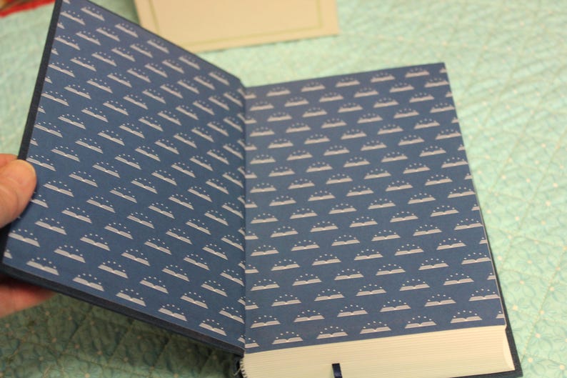 May include: An open hardcover book featuring dark blue endpapers with a repeating white pattern of open books topped with three small stars. The interior pages are white, and a thin dark blue ribbon bookmark is visible at the bottom edge.