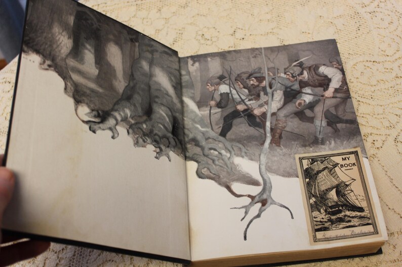 May include: Open book with a black and white illustration of a forest scene with figures in the background. The book is open to a page with a black and white illustration of a ship with the text "My Book" on the page.
