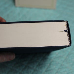 May include: A dark blue hardcover book viewed from the side, showing the cream-colored page edges. A small, dark blue fabric bookmark ribbon extends from the center of the page block, and the binding features a visible tear at the top edge.