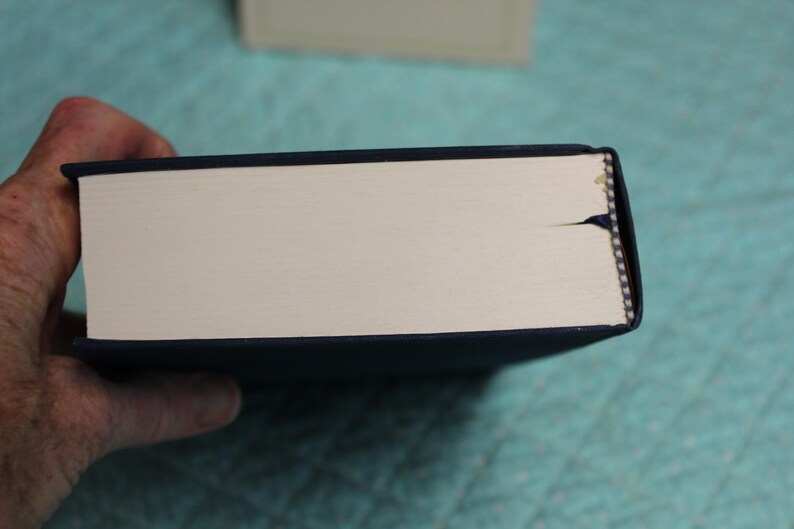 May include: A dark blue hardcover book viewed from the side, showing the cream-colored page edges. A small, dark blue fabric bookmark ribbon extends from the center of the page block, and the binding features a visible tear at the top edge.