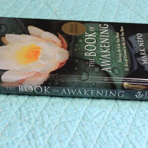 Może przedstawiać: Książka w twardej oprawie zatytułowana "The Book of Awakening" autorstwa Marka Nepo. Okładka przedstawia zbliżenie białej i ż&oacute;łtej lilii wodnej z kroplami wody. Książka jest wydaniem Deluxe i bestsellerem New York Times.