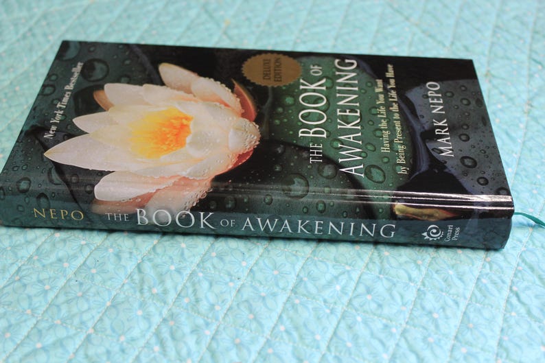 Może przedstawiać: Książka w twardej oprawie zatytułowana "The Book of Awakening" autorstwa Marka Nepo. Okładka przedstawia zbliżenie białej i ż&oacute;łtej lilii wodnej z kroplami wody. Książka jest wydaniem Deluxe i bestsellerem New York Times.