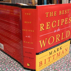 May include: A hardcover cookbook titled "The Best Recipes in the World" by Mark Bittman. The book has a red cover with yellow lettering and a yellow bottom section. The book's spine is also red with white text.