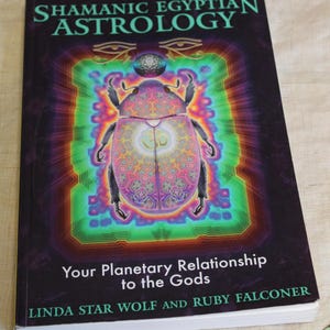 May include: A hardcover book titled "Shamanic Egyptian Astrology" with a colorful illustration of a scarab beetle. The book cover features the text "Your Planetary Relationship to the Gods" and the authors' names, Linda Star Wolf and Ruby Falconer.