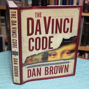 May include: A hardcover book titled The Da Vinci Code by Dan Brown, a special illustrated edition. The cover features a cream-colored background with dark red text and a torn paper graphic revealing a portion of the Mona Lisa painting's eyes.