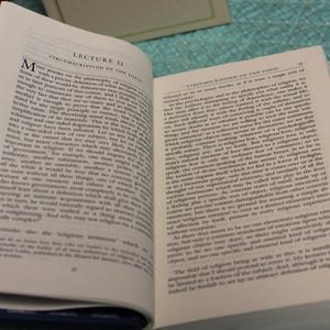 May include: An open hardcover book with white pages featuring black printed text. The left page is titled Lecture II Circumscription of the Topic. The book has a dark blue cover with a white pattern of small books and stars visible on the spine.