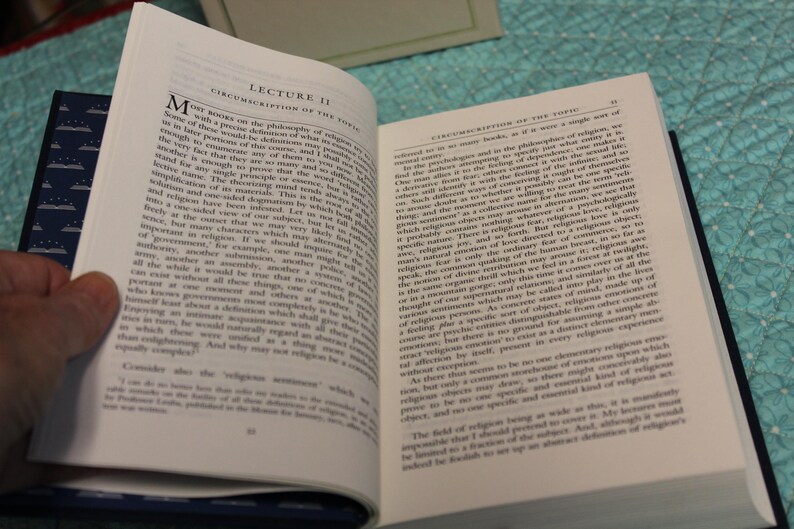 May include: An open hardcover book with white pages featuring black printed text. The left page is titled Lecture II Circumscription of the Topic. The book has a dark blue cover with a white pattern of small books and stars visible on the spine.